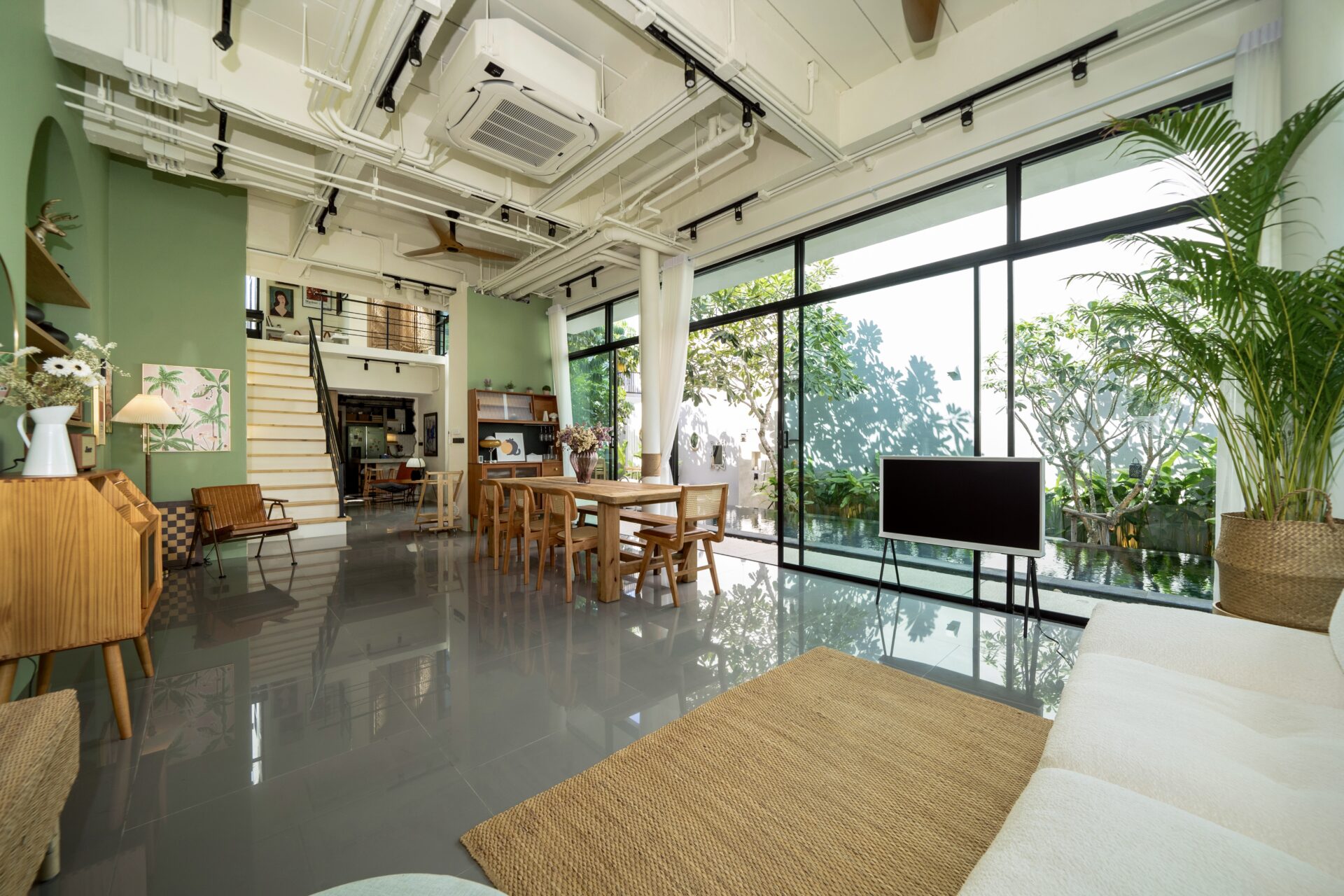 Open-concept living/dining area with large glass walls, wooden furniture, and lush indoor plants by a garden view.