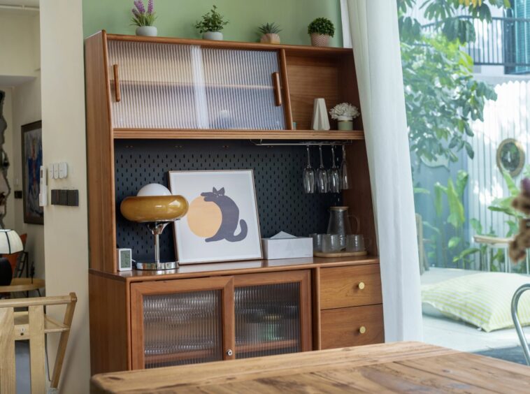 Wooden display cabinet with ribbed glass doors, plants on top, and a framed cat artwork on the lower shelf beside hanging wine glasses.
