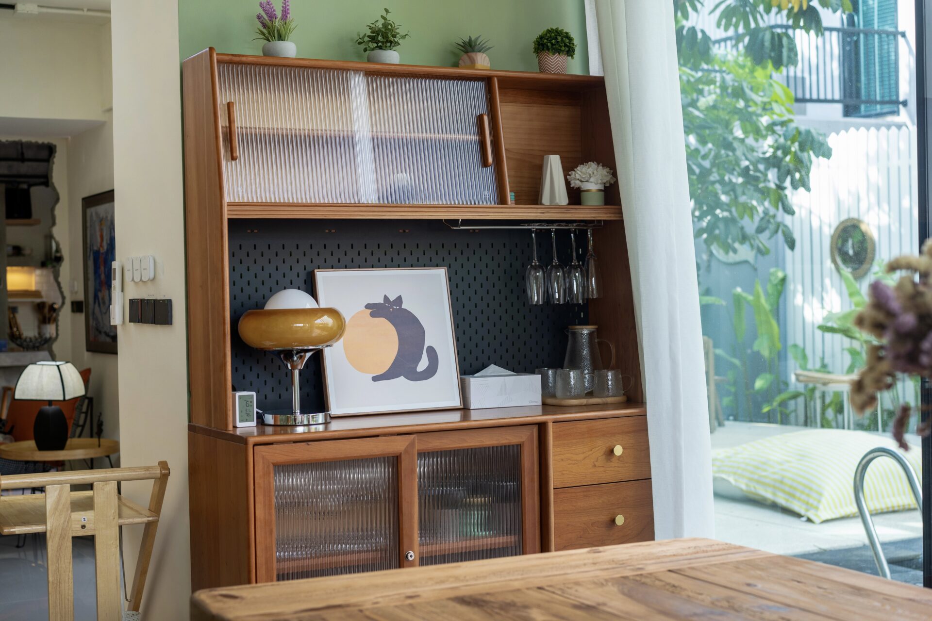 Wooden display cabinet with ribbed glass doors, plants on top, and a framed cat artwork on the lower shelf beside hanging wine glasses.