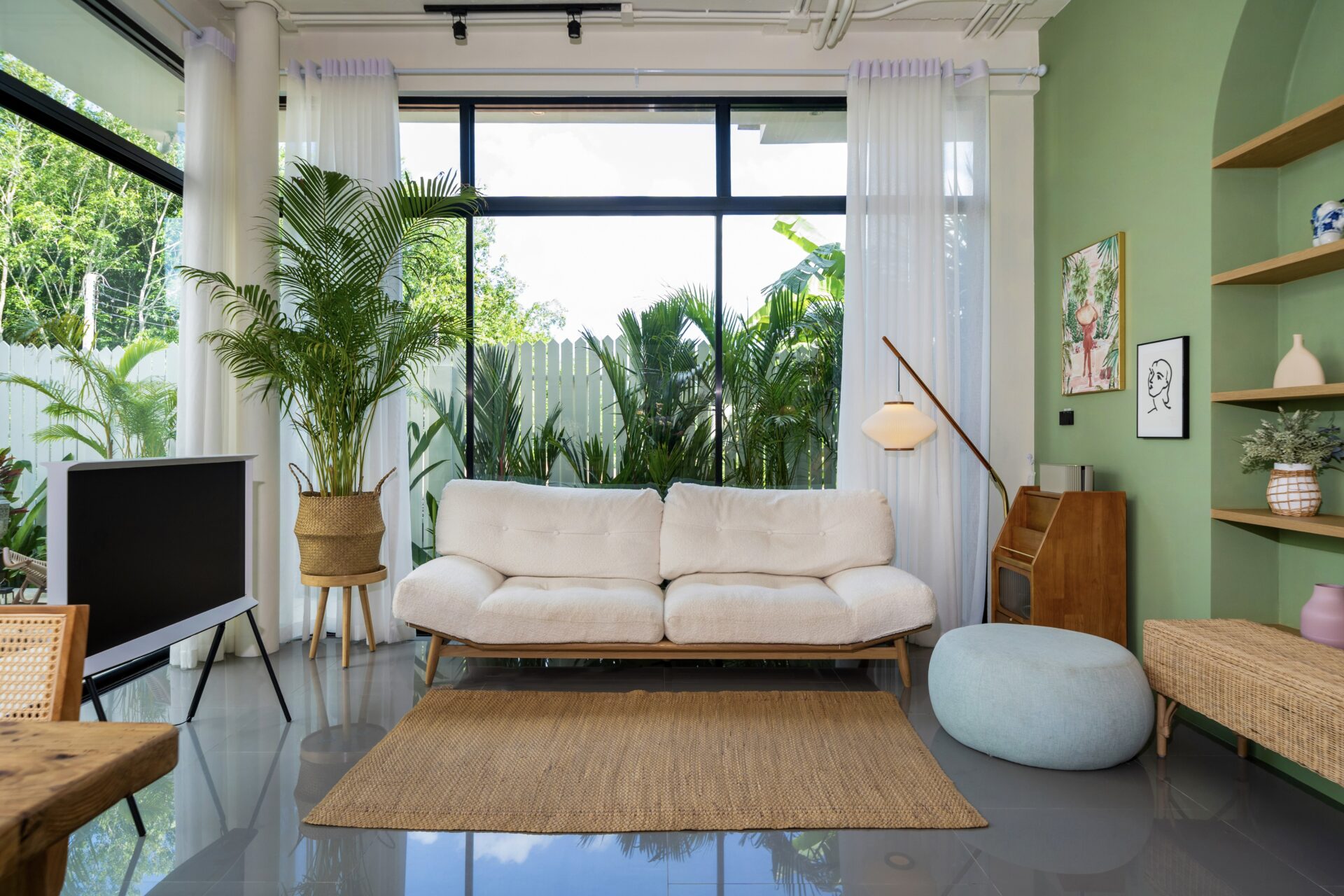 Bright living room with a white sofa, large glass windows, and lush indoor plants against a green wall.