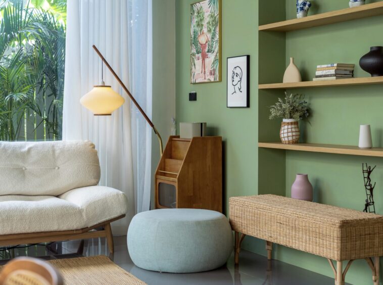 Bright green-walled living room with woven furniture, potted plants, and shelves displaying vases and books by a large window with sheer curtains.