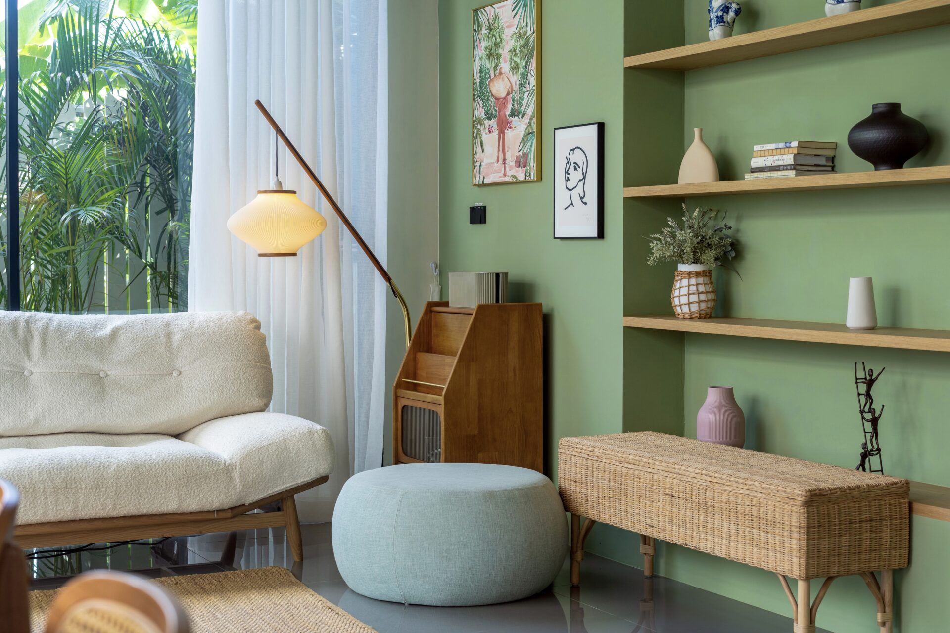 Bright green-walled living room with woven furniture, potted plants, and shelves displaying vases and books by a large window with sheer curtains.