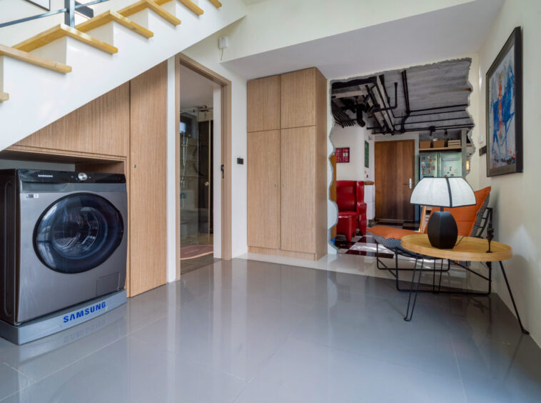Open-plan living area featuring a Samsung front-loading washing machine under wooden cabinetry, stairs overhead, and a seating nook with a round table lamp.