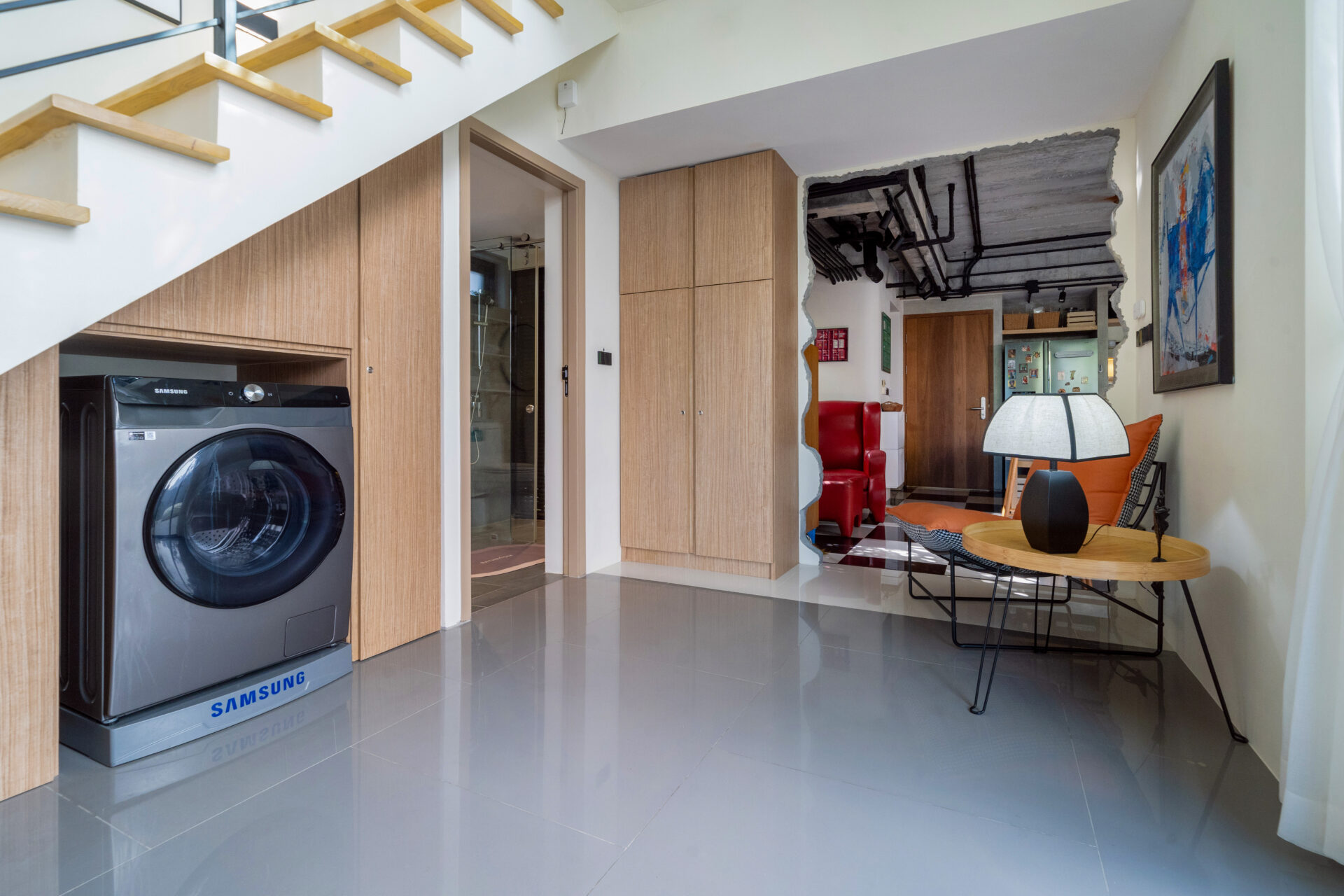 Open-plan living area featuring a Samsung front-loading washing machine under wooden cabinetry, stairs overhead, and a seating nook with a round table lamp.