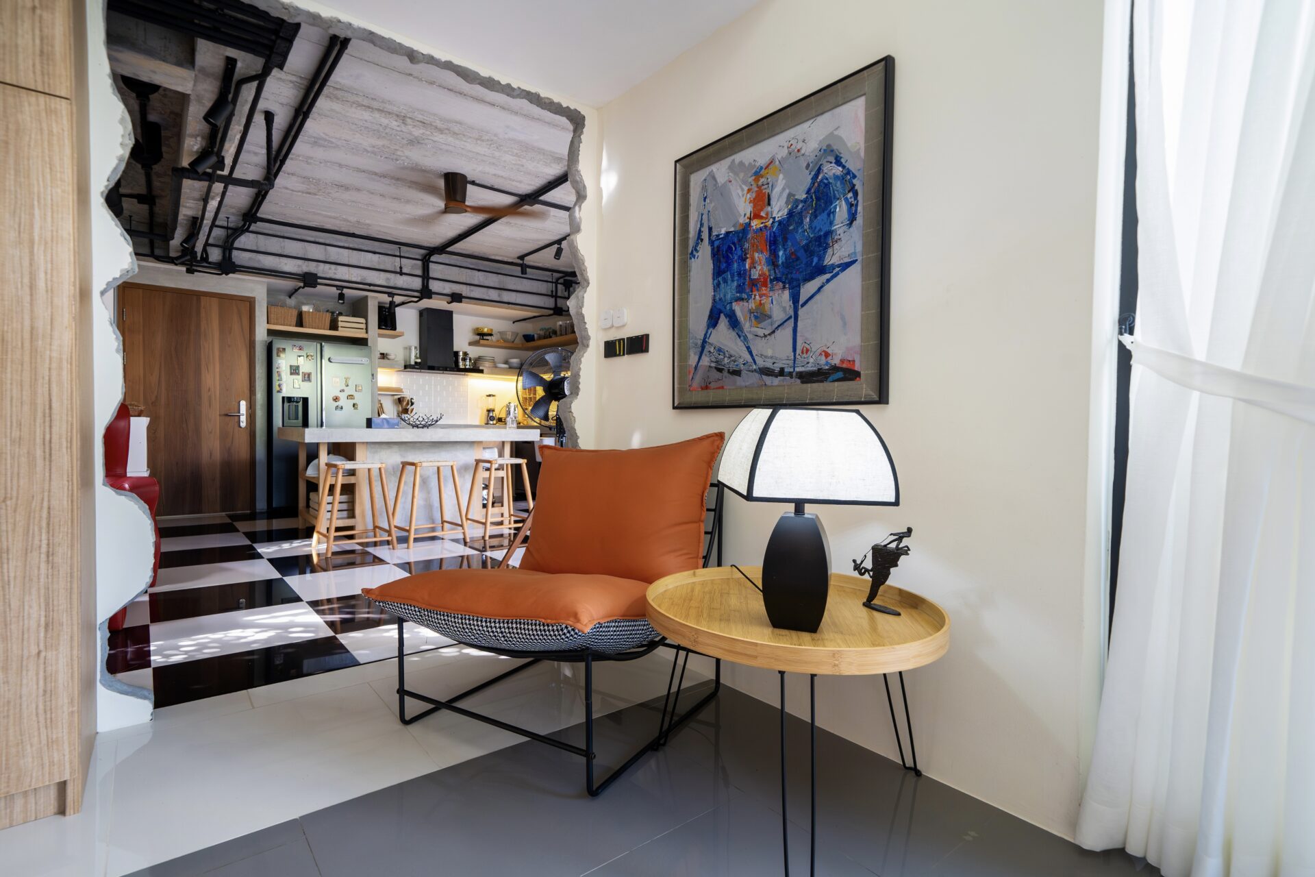 Open-plan living area with orange-cushioned chair and round wooden table facing a large abstract painting on the wall