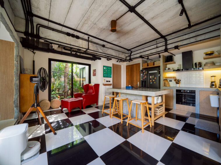 Industrial-style open-plan kitchen and living area with a black-and-white checkered floor, exposed ceiling pipes, and a red lounge chair near large glass doors opening to greenery.