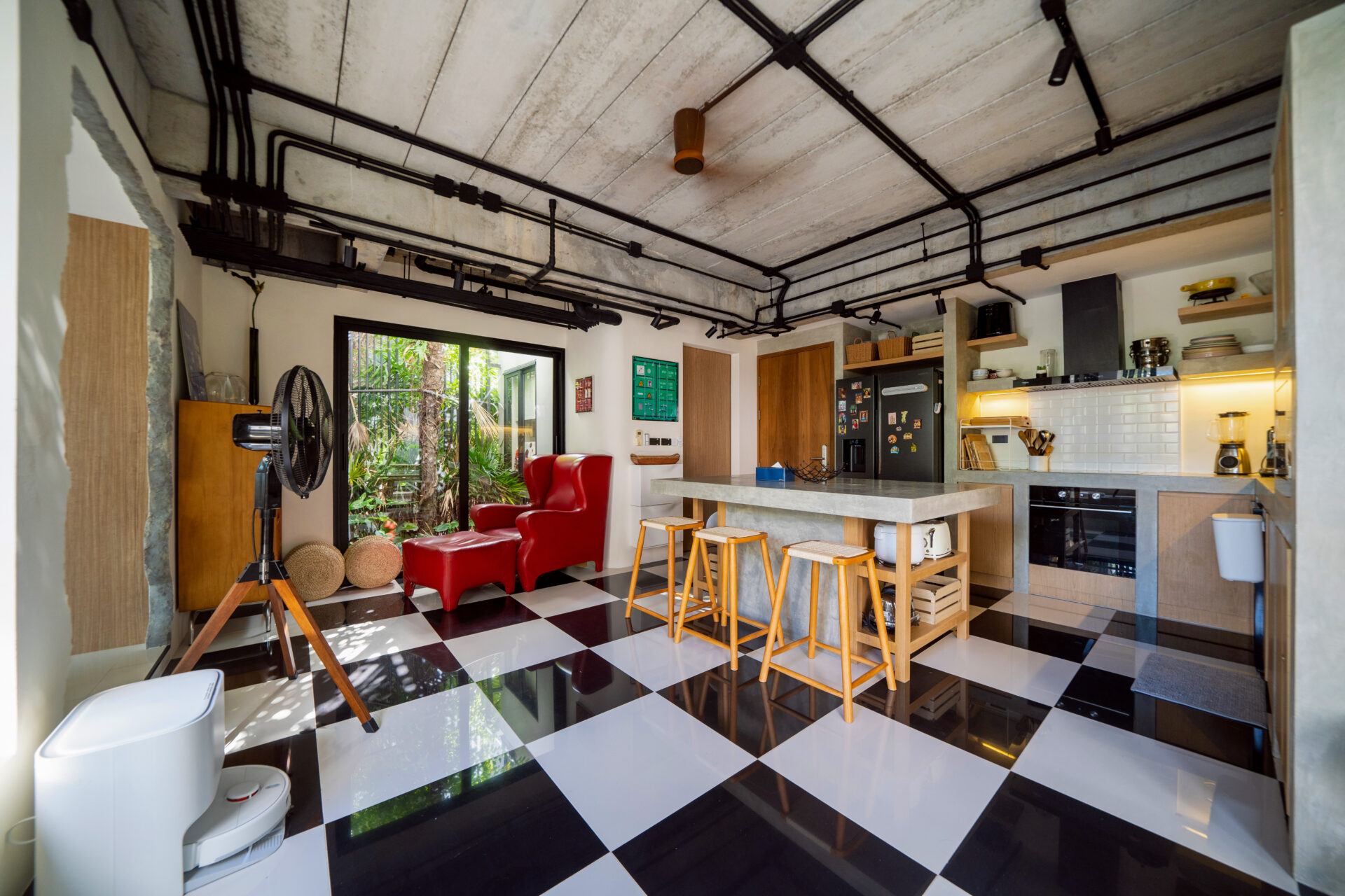 Industrial-style open-plan kitchen and living area with a black-and-white checkered floor, exposed ceiling pipes, and a red lounge chair near large glass doors opening to greenery.
