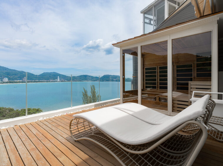 Sunlit wooden terrace with white lounge chairs overlooking calm blue ocean and distant hills through a glass railing