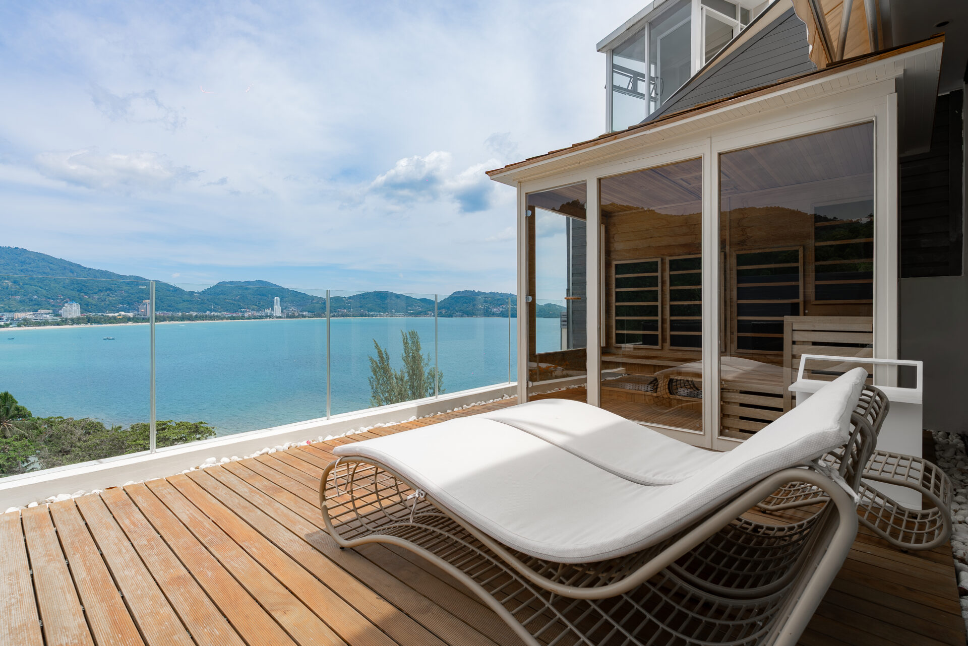 Sunlit wooden terrace with white lounge chairs overlooking calm blue ocean and distant hills through a glass railing
