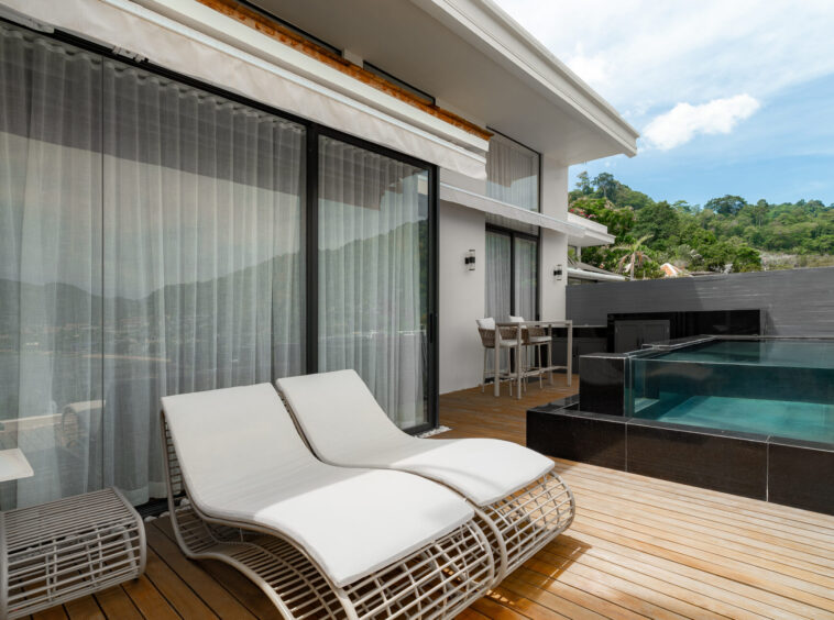 Outdoor poolside patio with white lounge chairs on a wooden deck beside glass sliding doors and sheer curtains behind them