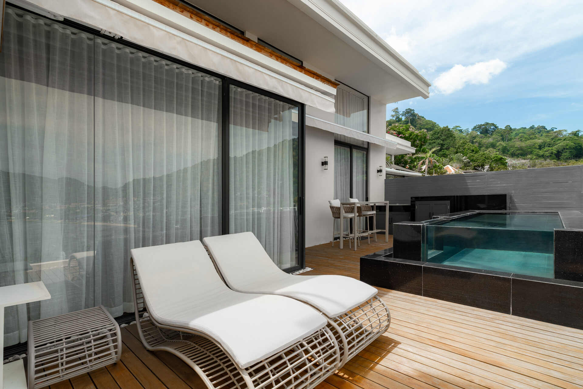 Outdoor poolside patio with white lounge chairs on a wooden deck beside glass sliding doors and sheer curtains behind them