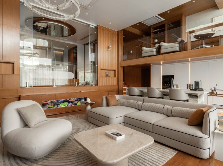 Luxurious open-plan living area with cream sectional sofa, marble coffee table, and warm wood paneling; glass mezzanine in background.