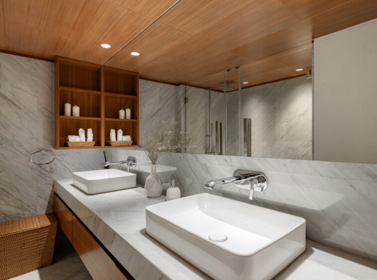 Modern bathroom with two rectangular white sinks on a marble countertop and a wooden cabinet above, marble walls, and a wicker laundry basket in the corner