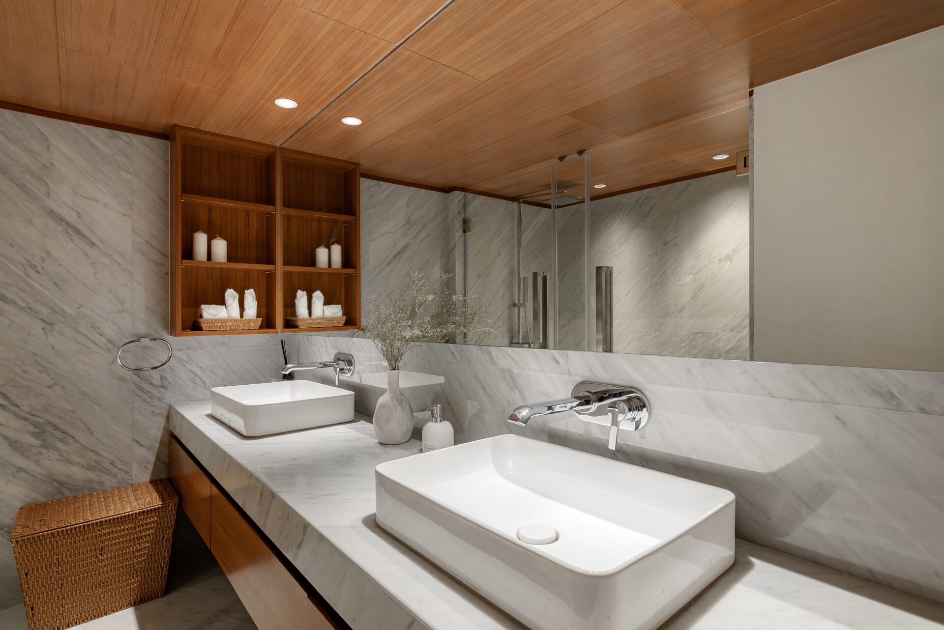 Modern bathroom with two rectangular white sinks on a marble countertop and a wooden cabinet above, marble walls, and a wicker laundry basket in the corner