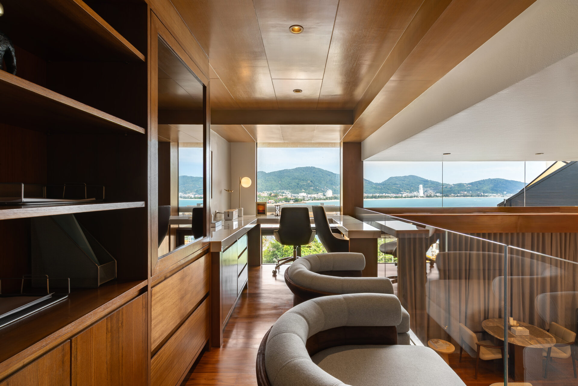 Modern home office with curved gray seating, glass railing, and built-in wooden shelves overlooking a bay and hills outside.