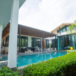 Resort-style villa with a rectangular pool, outdoor seating under a covered patio, and tropical hedges along the edge.
