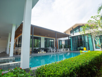 Resort-style villa with a rectangular pool, outdoor seating under a covered patio, and tropical hedges along the edge.