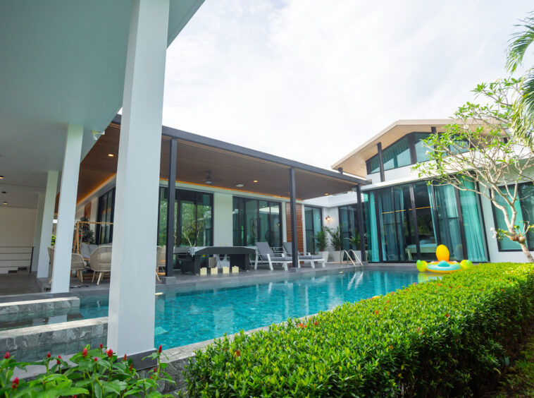 Resort-style villa with a rectangular pool, outdoor seating under a covered patio, and tropical hedges along the edge.
