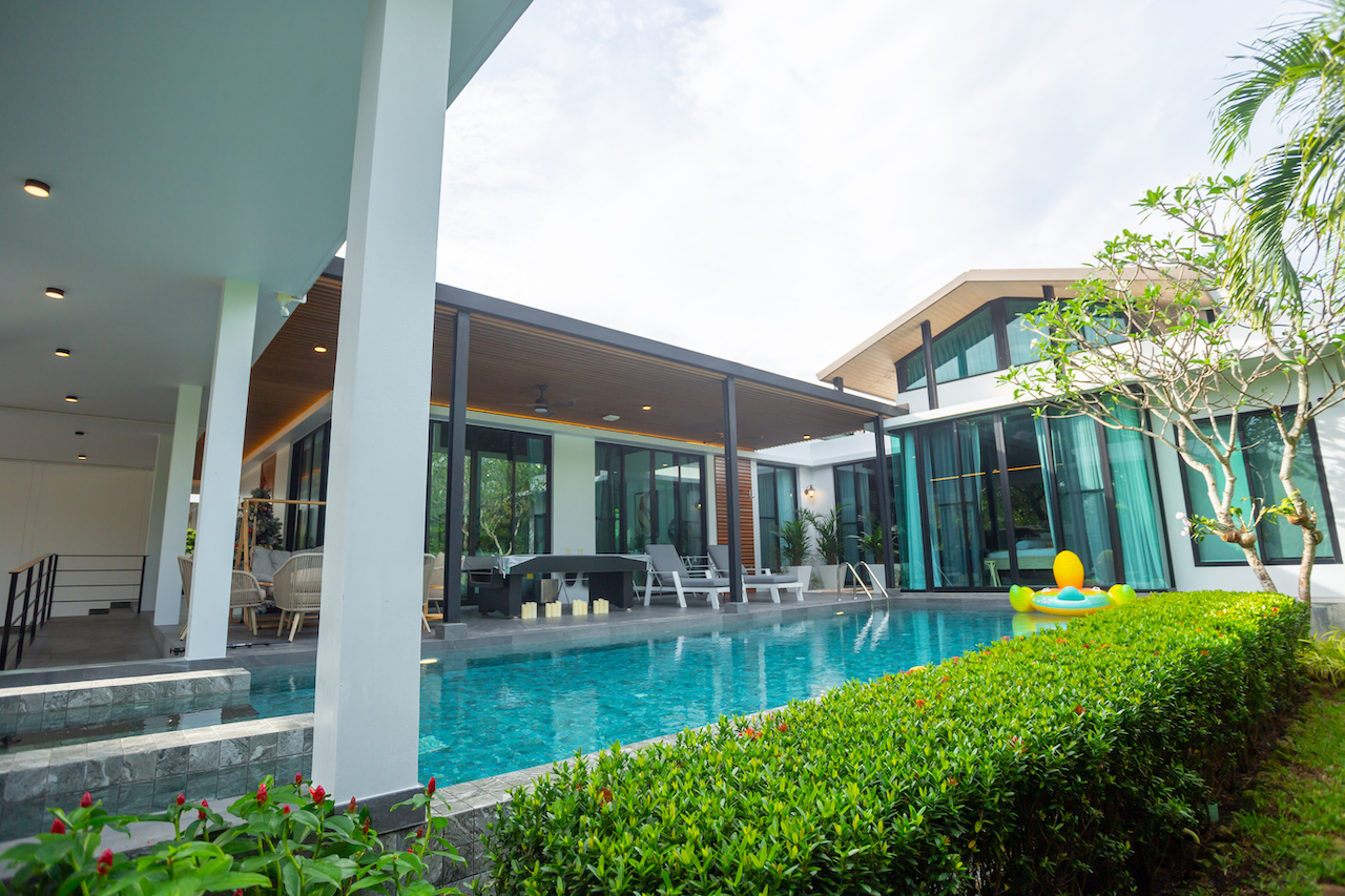 Resort-style villa with a rectangular pool, outdoor seating under a covered patio, and tropical hedges along the edge.