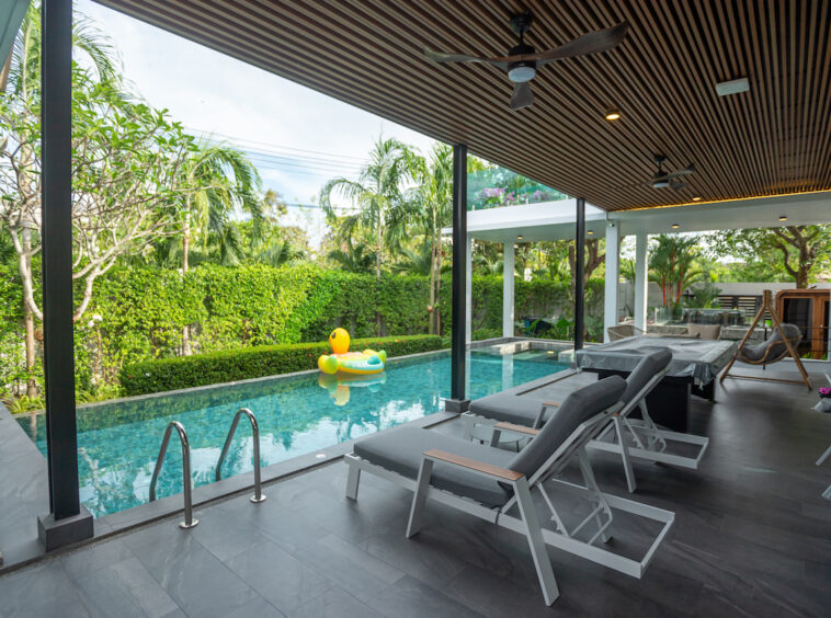 Covered poolside patio with row of gray lounge chairs facing a rectangular turquoise pool; inflatable yellow duck floats in the water, and lush hedges surround the area.