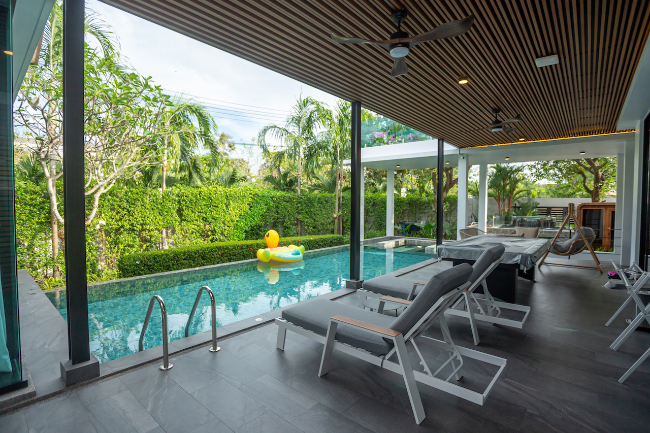 Covered poolside patio with row of gray lounge chairs facing a rectangular turquoise pool; inflatable yellow duck floats in the water, and lush hedges surround the area.