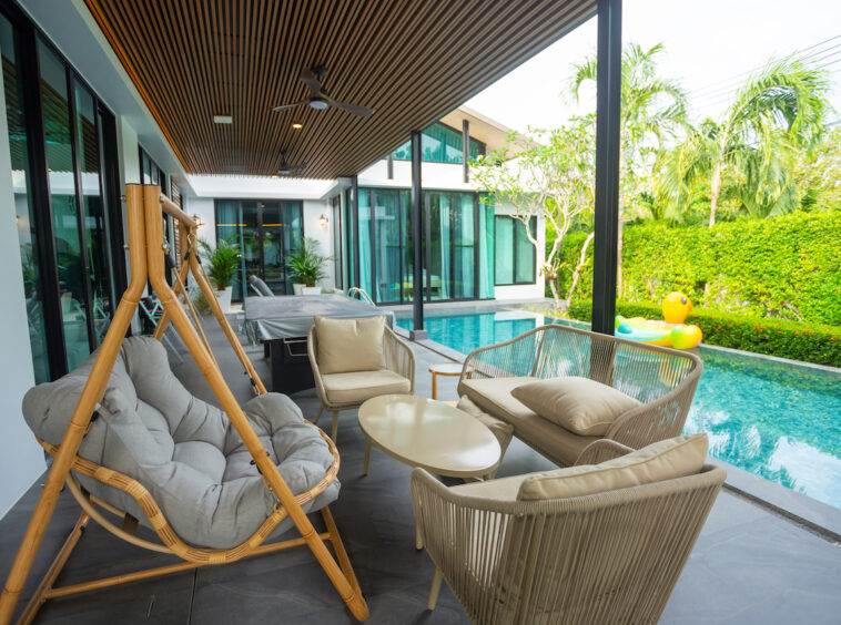 Covered patio with beige cushioned wicker seating around a white coffee table beside a narrow pool.