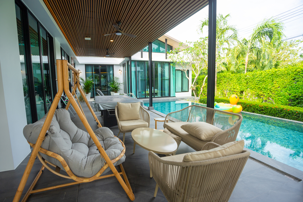 Covered patio with beige cushioned wicker seating around a white coffee table beside a narrow pool.