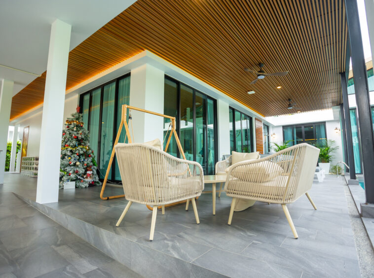 Outdoor lounge area with beige wicker chairs around a glass table under a wooden slatted ceiling, near a Christmas tree