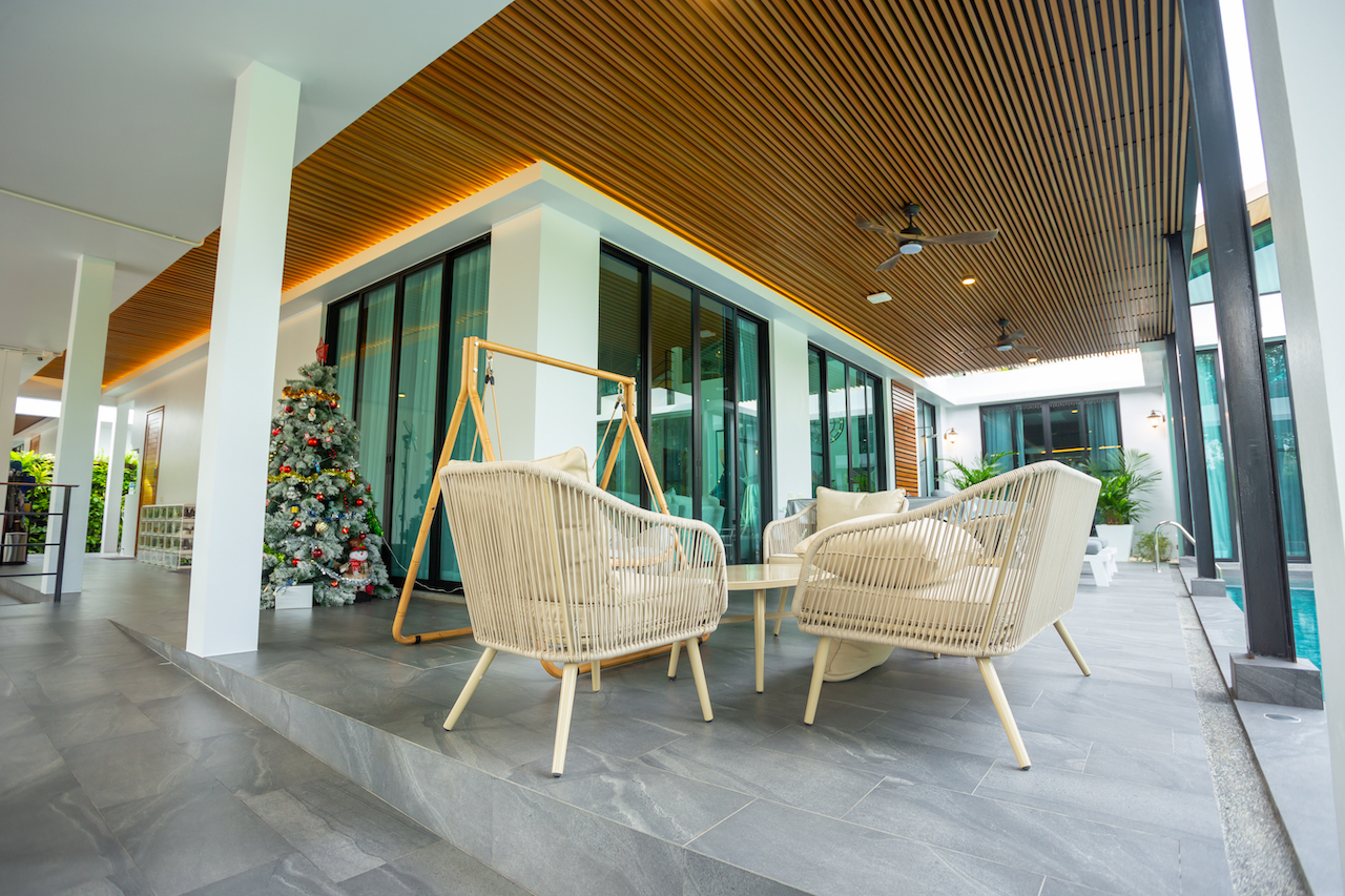 Outdoor lounge area with beige wicker chairs around a glass table under a wooden slatted ceiling, near a Christmas tree