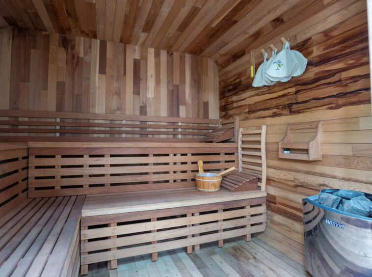 Wooden sauna interior with multi‑level benches, heater with rocks, and a wooden bucket with a ladle on the bench.
