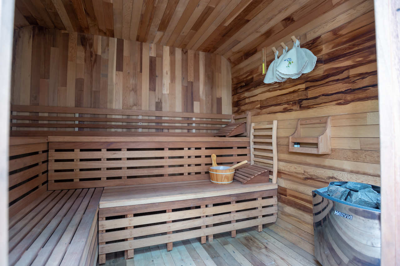 Wooden sauna interior with multi‑level benches, heater with rocks, and a wooden bucket with a ladle on the bench.