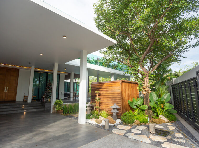 Modern covered patio with white columns, dark tile flooring, and a wooden door at the left; garden area with rocks and greenery in the center-right, and a curved wooden shed behind.