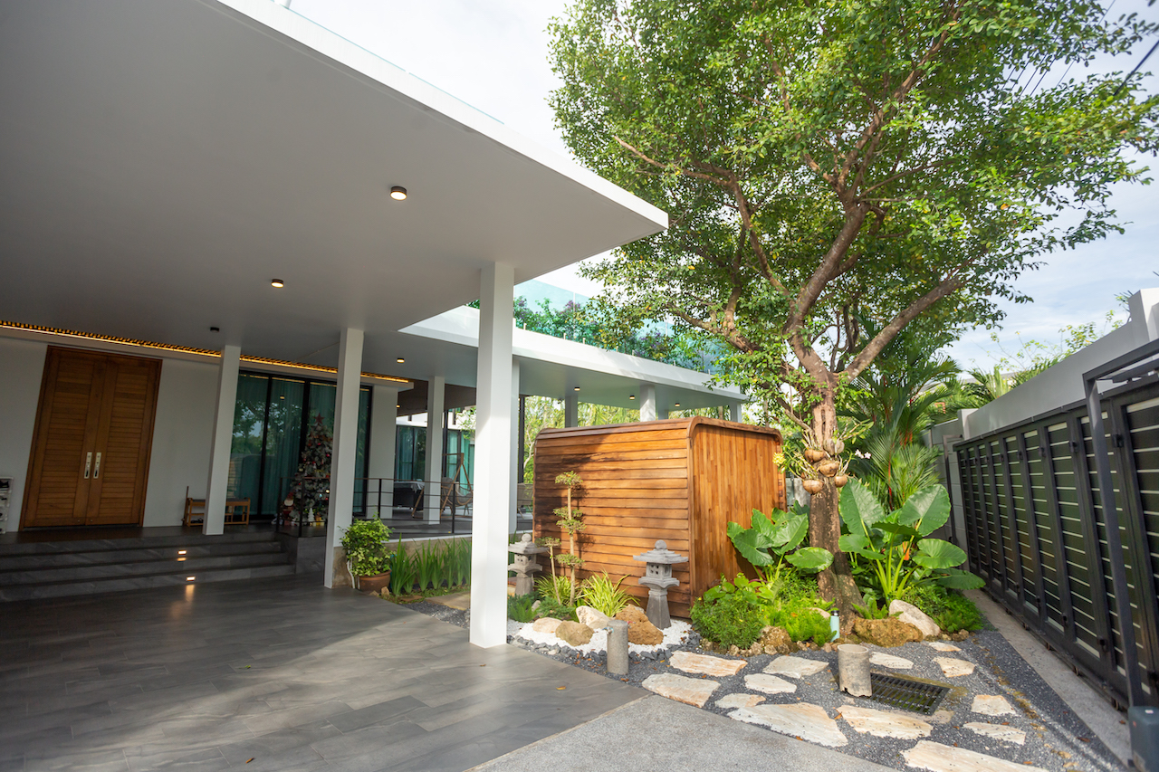 Modern covered patio with white columns, dark tile flooring, and a wooden door at the left; garden area with rocks and greenery in the center-right, and a curved wooden shed behind.