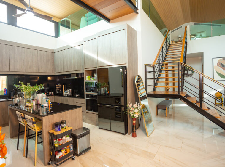 Open-plan kitchen with a large dark island, grey wood cabinets, built-in ovens, and a glossy tiled floor, adjacent to a staircase with a metal railing leading to a mezzanine.