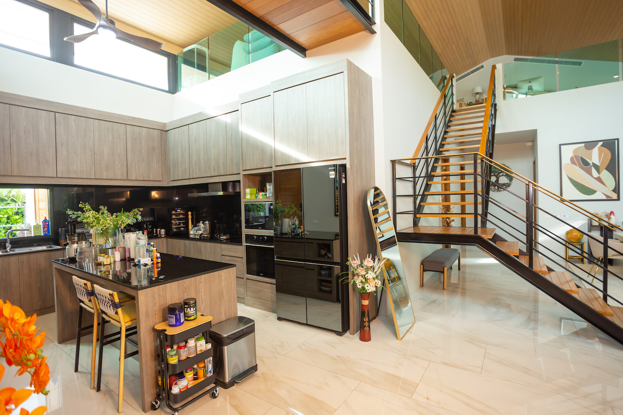 Open-plan kitchen with a large dark island, grey wood cabinets, built-in ovens, and a glossy tiled floor, adjacent to a staircase with a metal railing leading to a mezzanine.