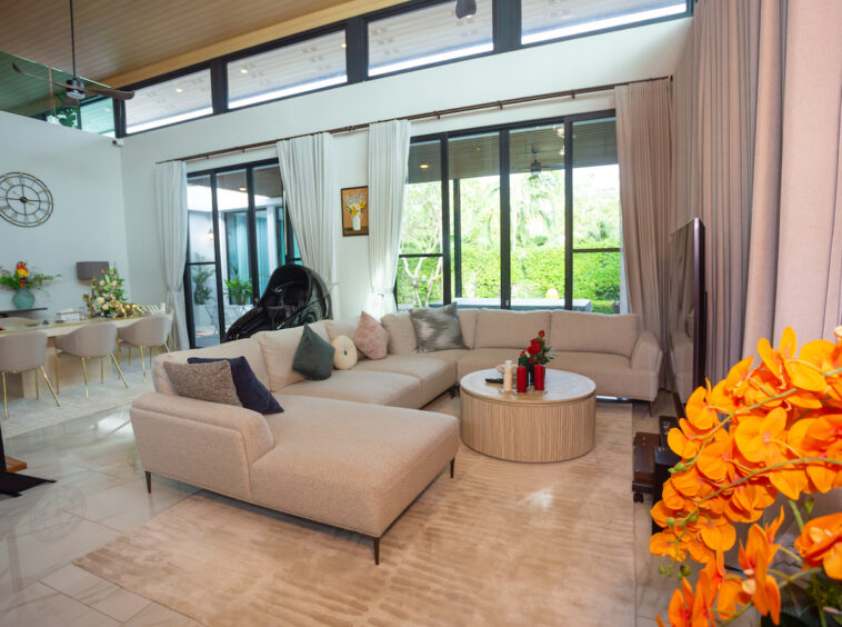 Bright open-plan living room with a beige sectional, round coffee table, and orange flowers in the foreground; large windows with cream curtains overlook a garden area.