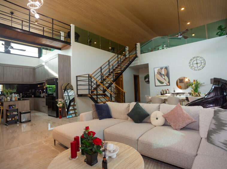 Open-plan living area with a large beige sectional, round coffee table, decorative cushions, and a metal staircase to a mezzanine.