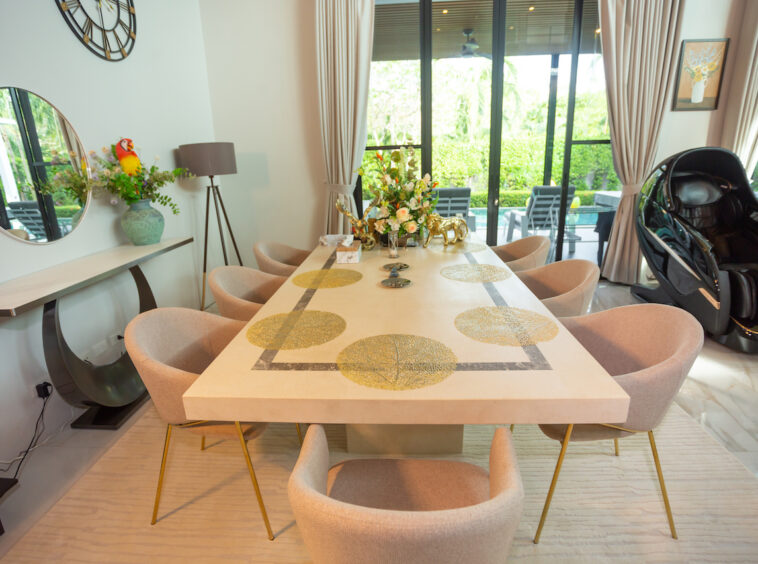 Long cream dining table with gold circular inlays surrounded by beige chairs in a bright room with large glass doors on one side, and a massage chair nearby.