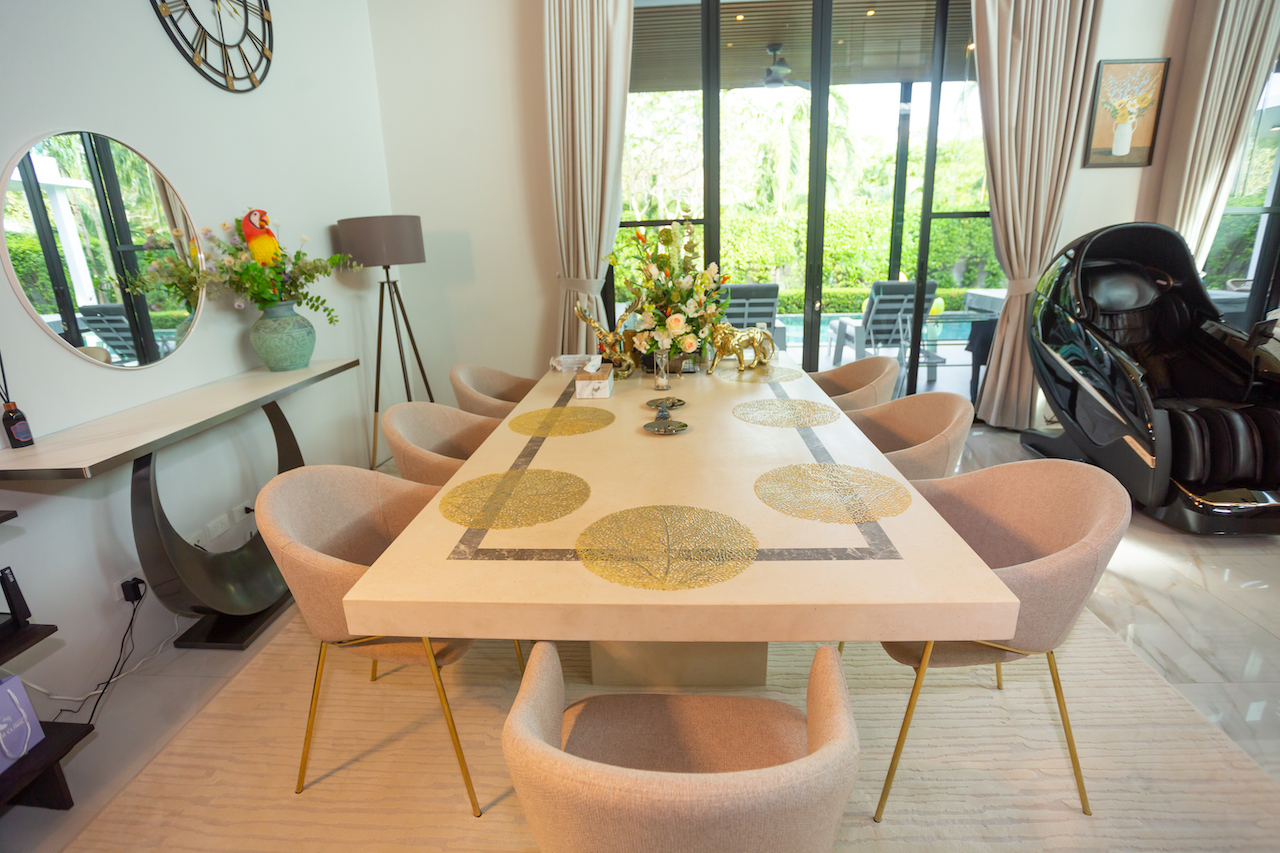 Long cream dining table with gold circular inlays surrounded by beige chairs in a bright room with large glass doors on one side, and a massage chair nearby.