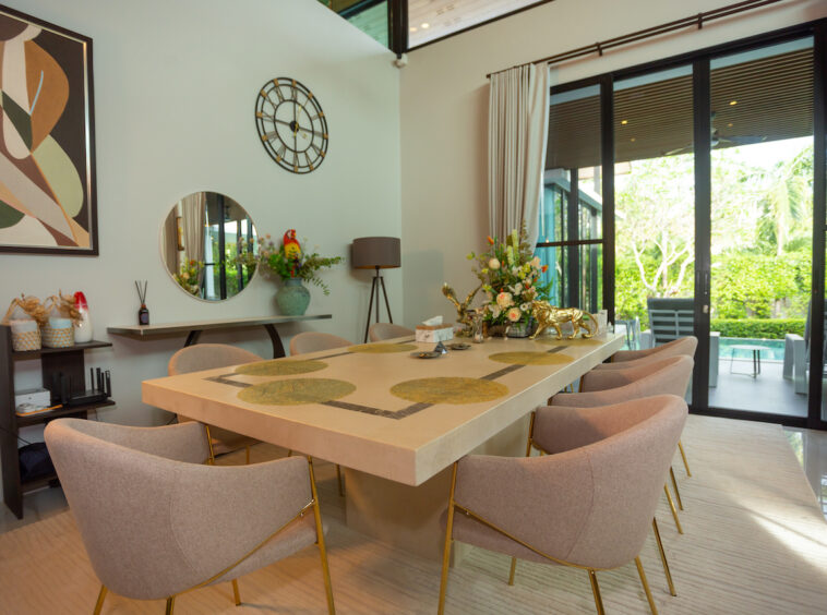 Elegant dining room with a long light-colored table, beige upholstered chairs, and a floral centerpiece by large glass doors to a patio.