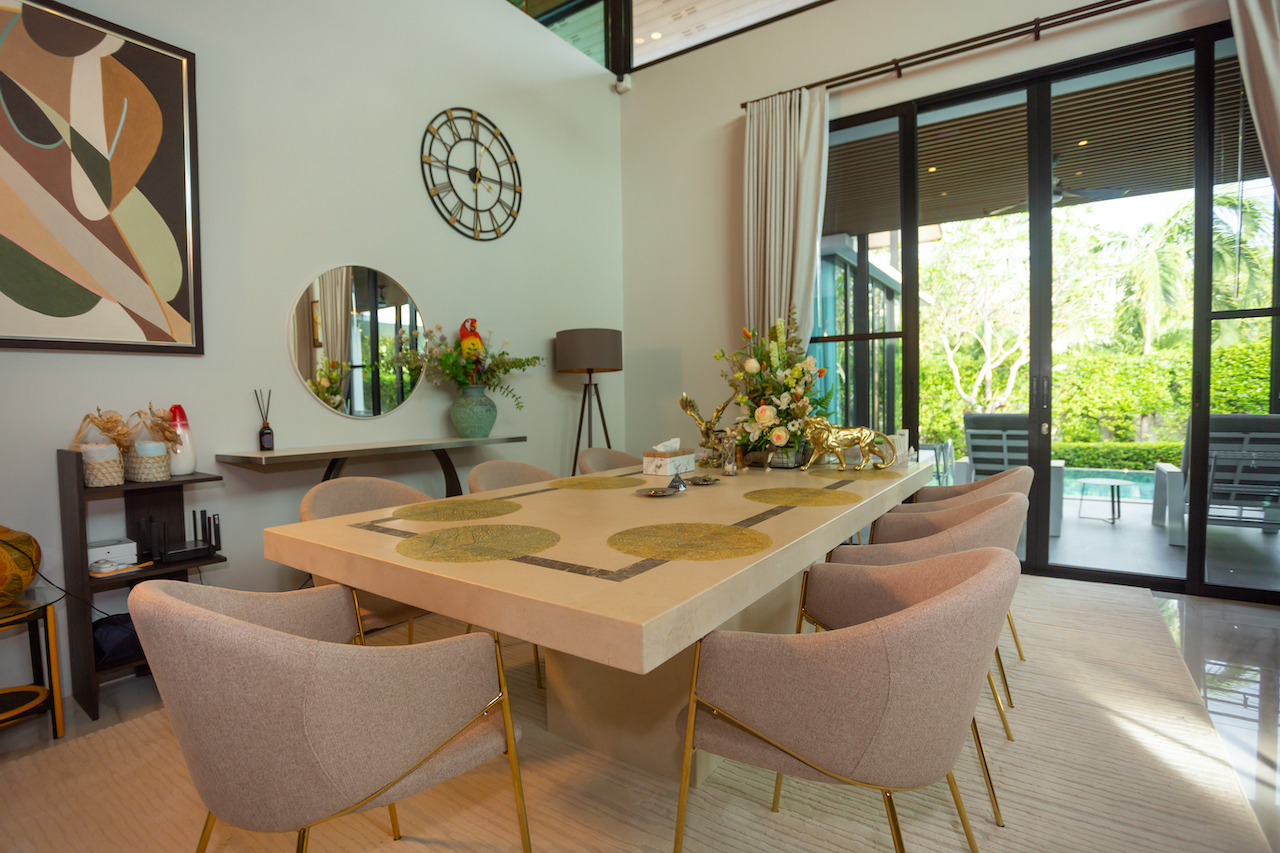 Elegant dining room with a long light-colored table, beige upholstered chairs, and a floral centerpiece by large glass doors to a patio.