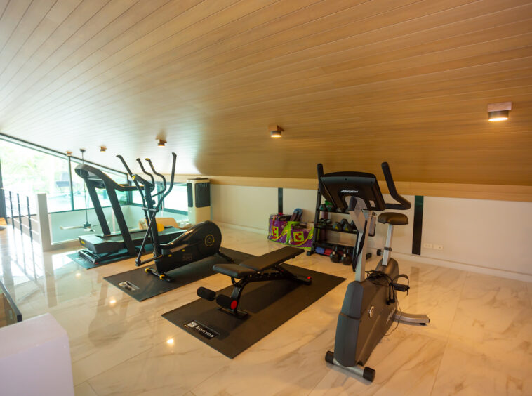 Home gym with treadmill, elliptical, stationary bike on black mats under a wooden sloped ceiling, bright loft setting.