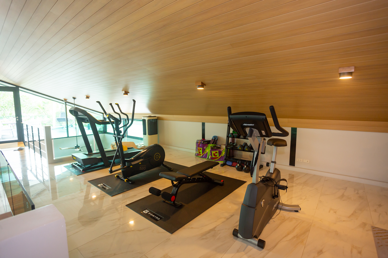 Home gym with treadmill, elliptical, stationary bike on black mats under a wooden sloped ceiling, bright loft setting.