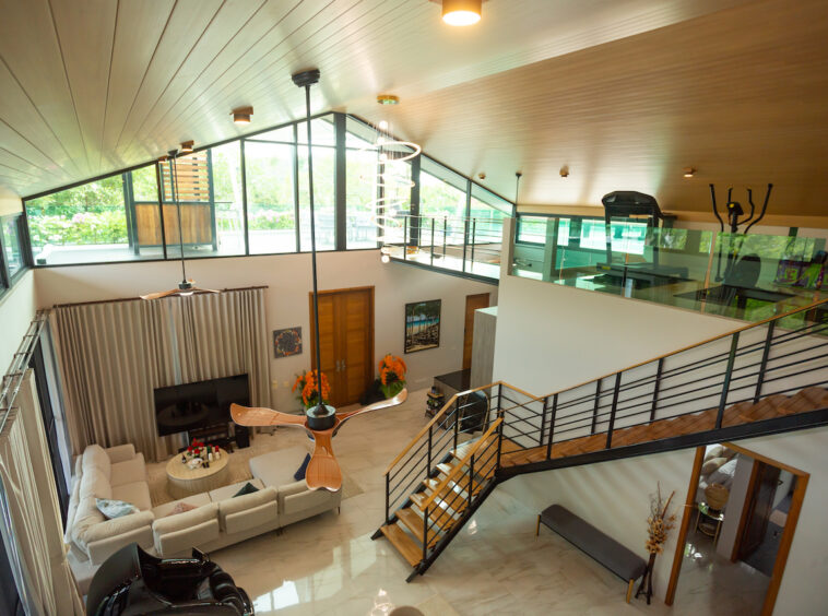 Two-story living room with a lofted gym area, large windows, and a curved wooden ceiling; beige sectional sofa and round coffee table beneath a modern pendant light.