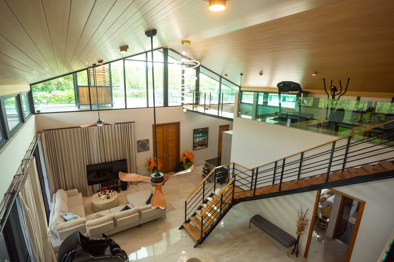 Two-story living room with a lofted gym area, large windows, and a curved wooden ceiling; beige sectional sofa and round coffee table beneath a modern pendant light.