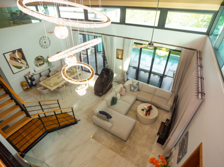 Spacious modern living room seen from above with a beige sectional sofa, circular chandelier, and glass doors to a pool outside.