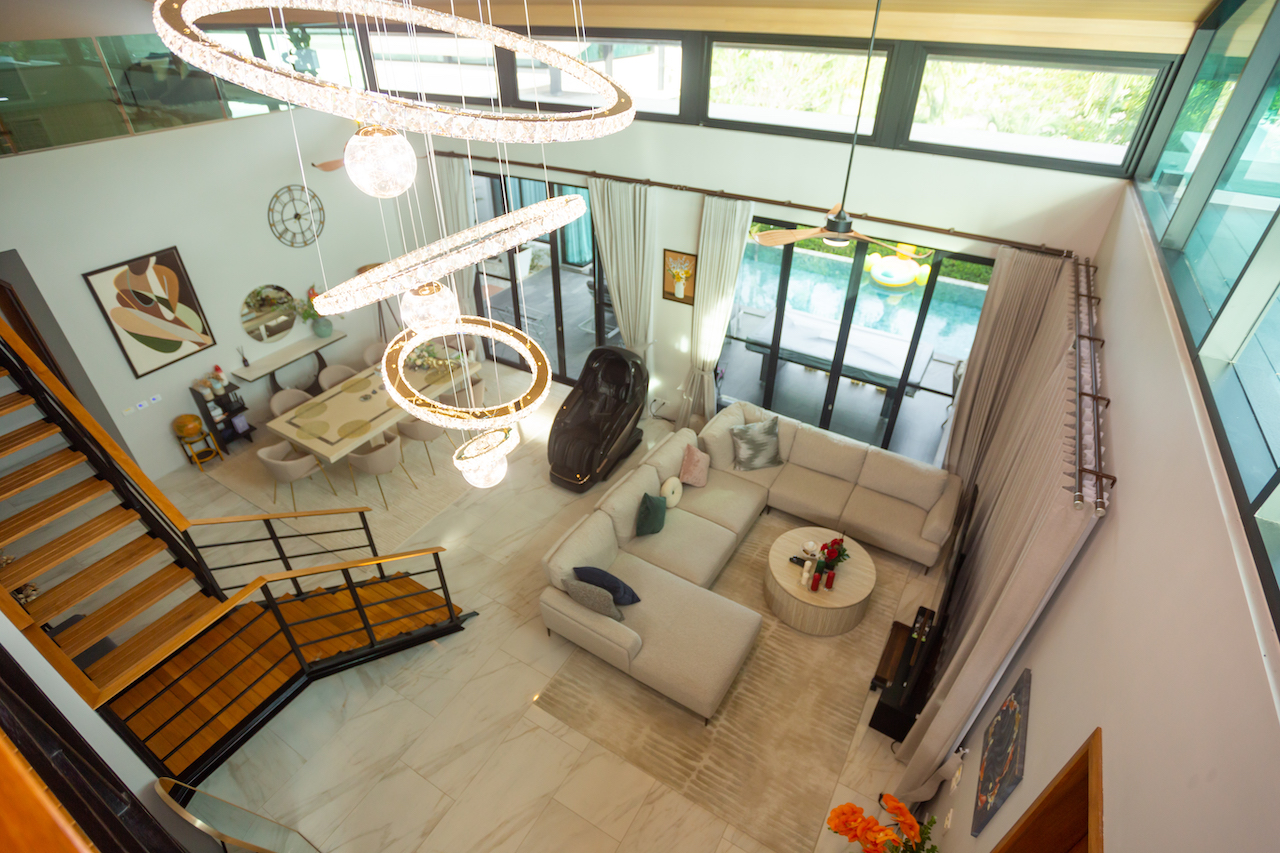 Spacious modern living room seen from above with a beige sectional sofa, circular chandelier, and glass doors to a pool outside.