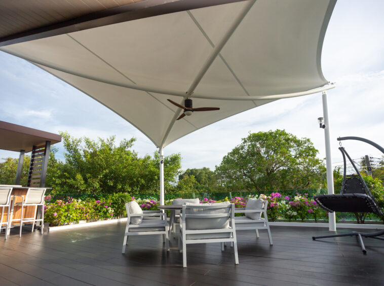 Outdoor patio with white furniture under a fabric shade sail and a ceiling fan, overlooking a garden with pink flowers.