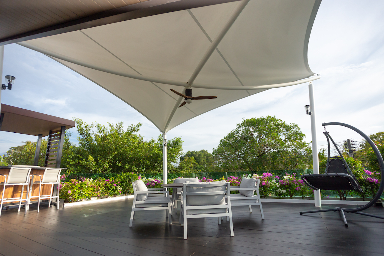 Outdoor patio with white furniture under a fabric shade sail and a ceiling fan, overlooking a garden with pink flowers.