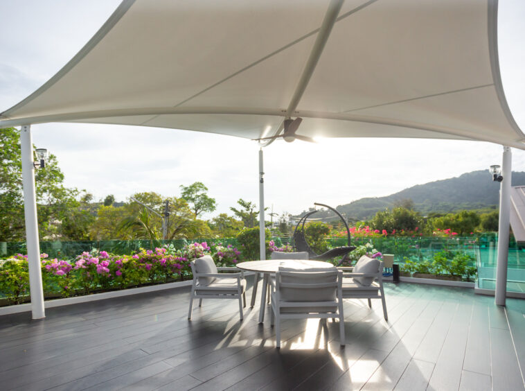 Outdoor terrace with a large curved white shade, round table and four chairs, flowering garden and hills in the background.